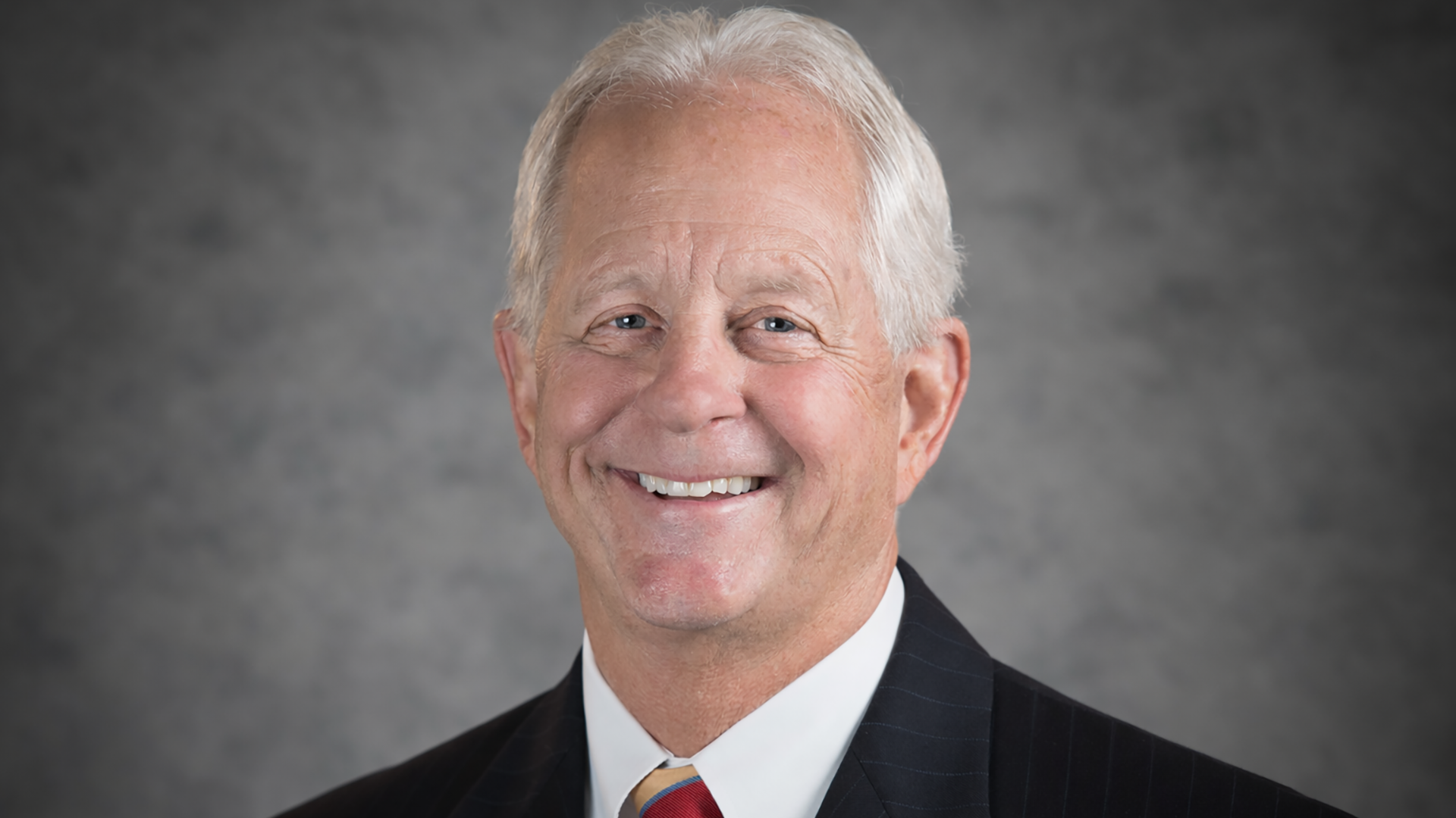 A man with white hair and black suit smiling